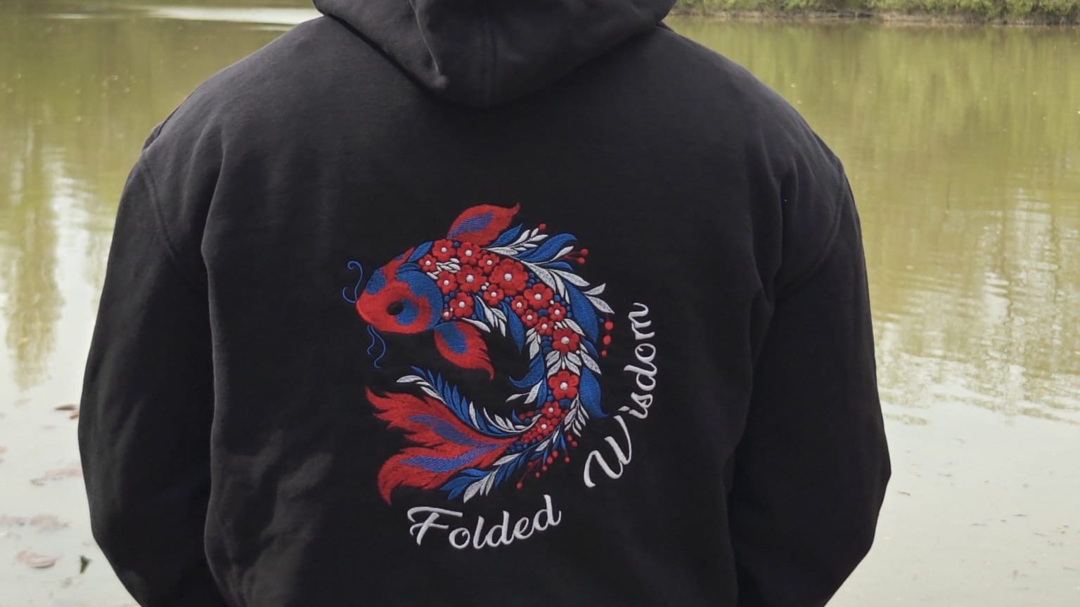 Back of a black Folded Wisdom hoodie with an embroidered koi, by a lake